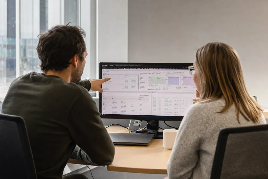 Zwei Mitarbeitende betrachten gemeinsam ein Monitoring-Dashboard auf einem Bildschirm.